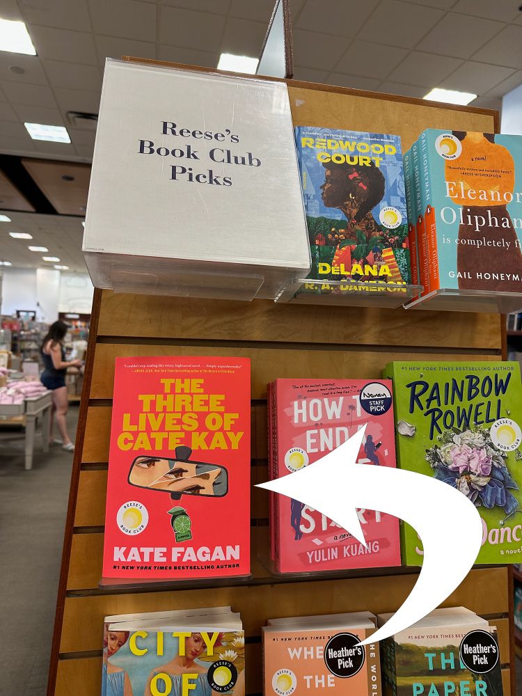 A display of paperback books at an Indigo book store featuring the books from Reese's Book Club. An arrow points to the book The Three Lives Of Cate Kay by Kate Fagan, which is a red cover with an illustration of broken trio of woman's eyes in a rearview car mirror. 
