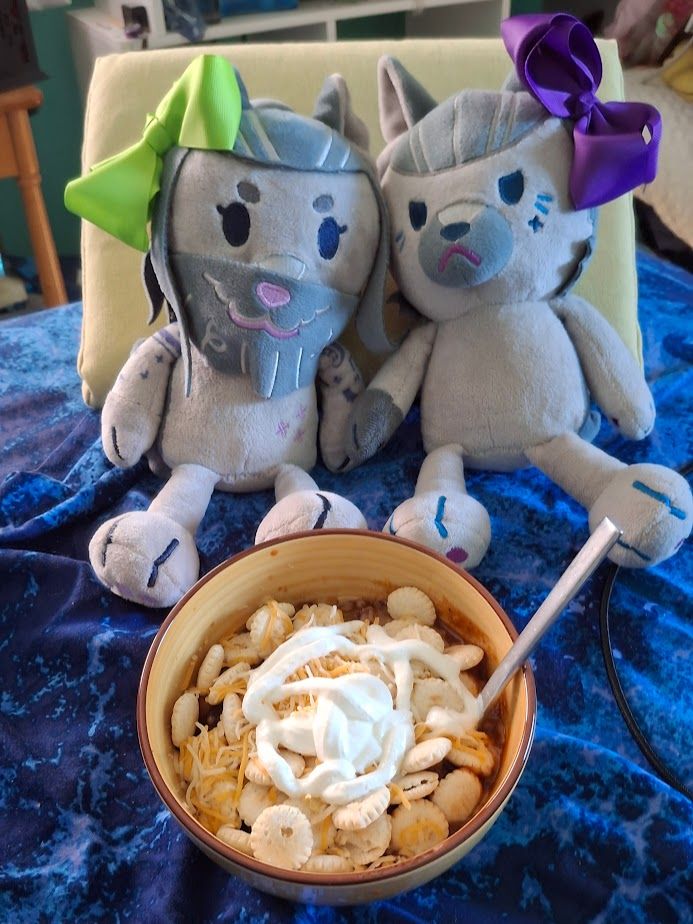 Doggy Izzy and Wolf Ed plushes posing in front of a bowl of chili topped with oyster crackers and shredded cheese and sour cream