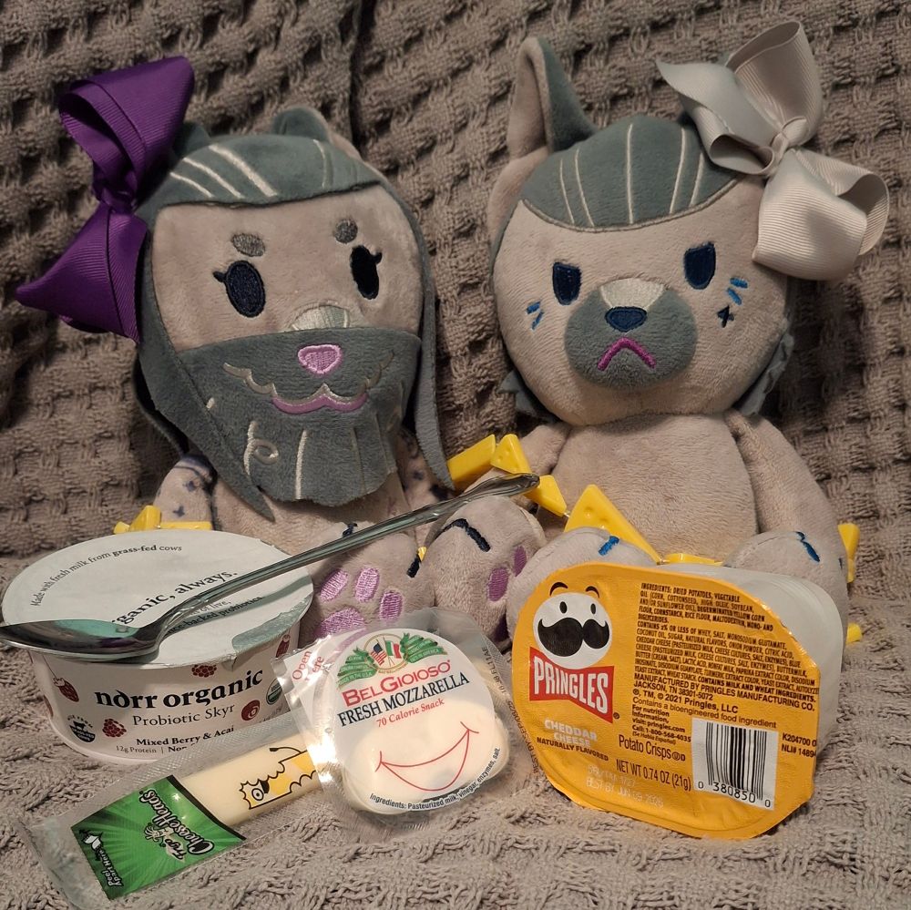 Doggy Izzy and Wolf Ed plushes sitting with some string cheese, fresh mozzarella cheese, skyr, and cheddar cheese Pringles