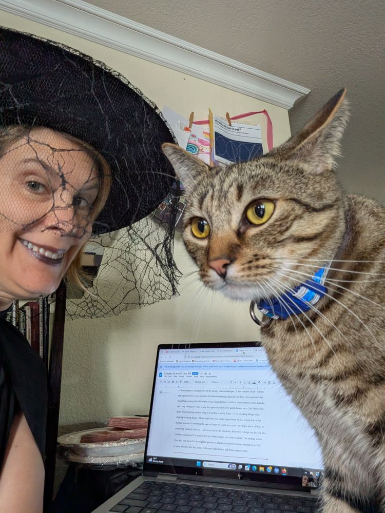 Woman in black witch's costume and gloves with her computer and a gold-eyed tabby cat standing in the way.