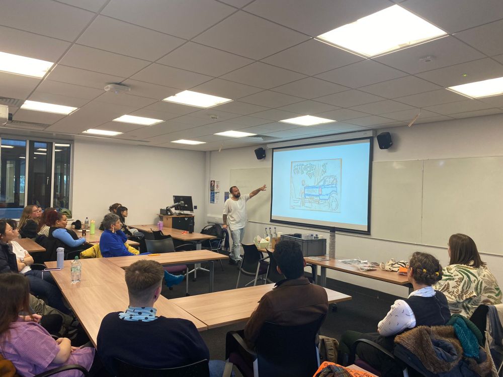 A presenter points to the smartboard, which shows an illustration of the back of a jeep or truck with a figure standing beside it and a tree behind. 14 audience members are shows, sitting around tables in a horseshoe layout, all attentively looking towards the presenter and screen.