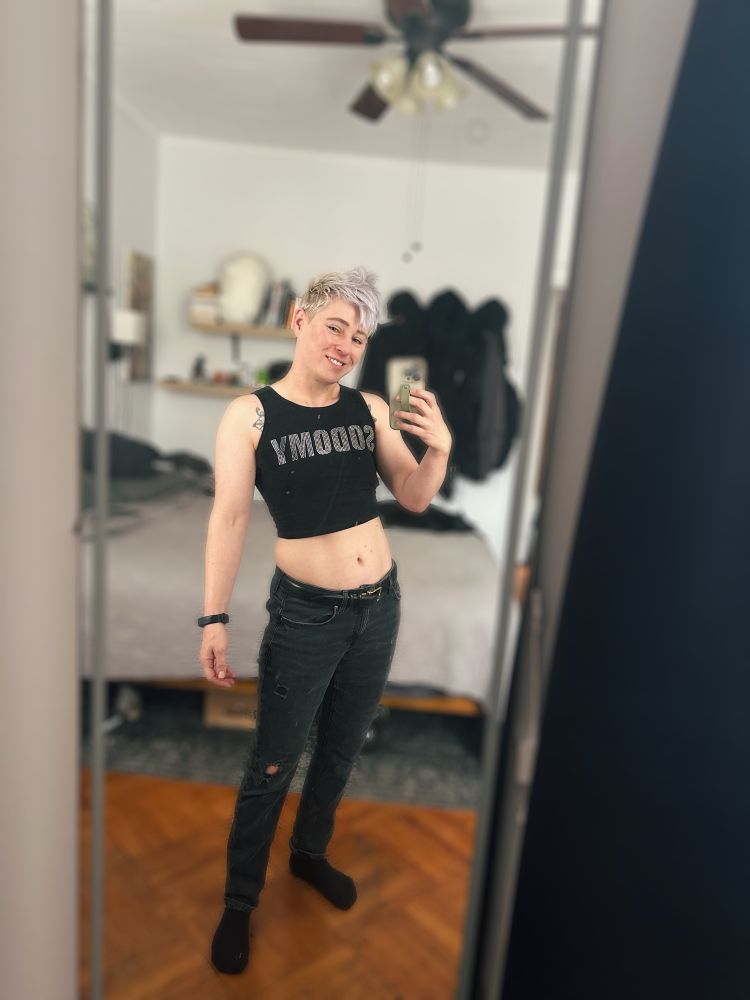 Jaq taking a mirror selfie, smiling, wearing black jeans and a black crop top with SODOMY written in rhinestones on it.