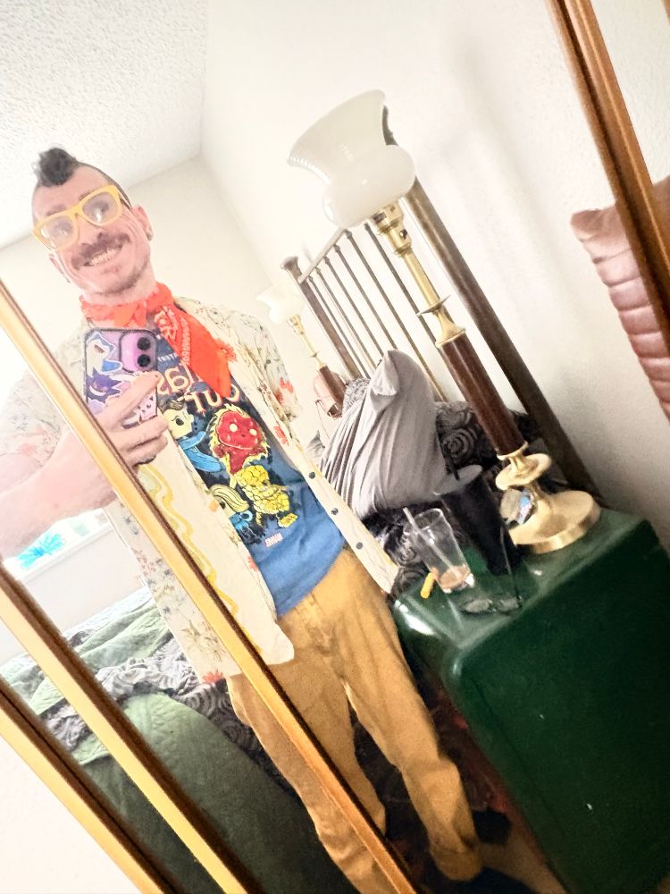 Brad in front of a mirror wearing a fantastic four shirt, floral button down, orange bandana and yellow jeans 