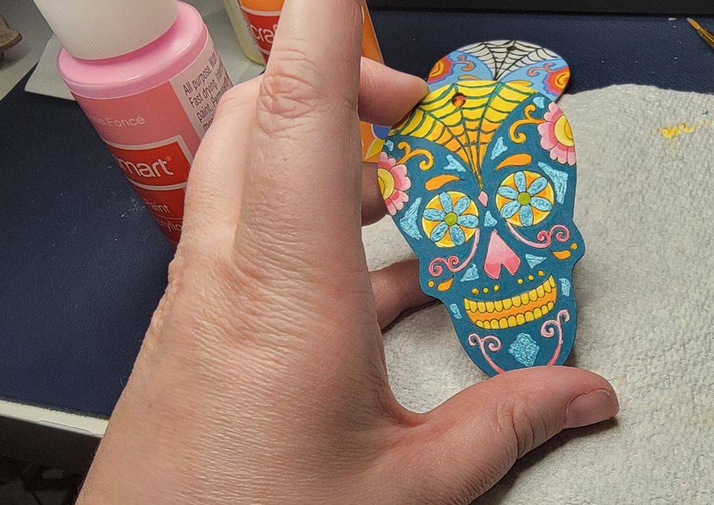 A paint-stained hand holds up a sugar skull painted in turquoise, yellow, orange and pink. There are acrylic paints in the background. 