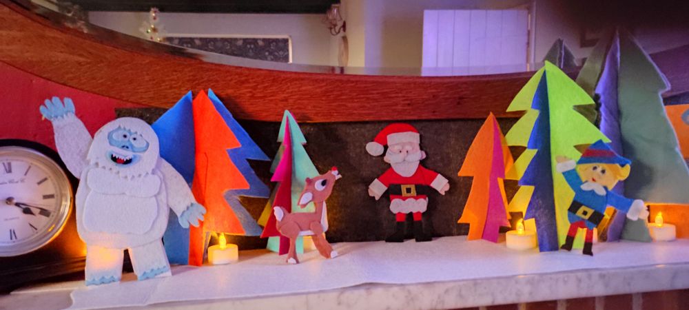 A mantle piece with a scene made with felt arranged on it. An Abominable Snowman, Rudolph, Santa and Hermey the Misfit Elf from the Rankin and Bass movies are stood amongst brightly coloured Christmas trees.