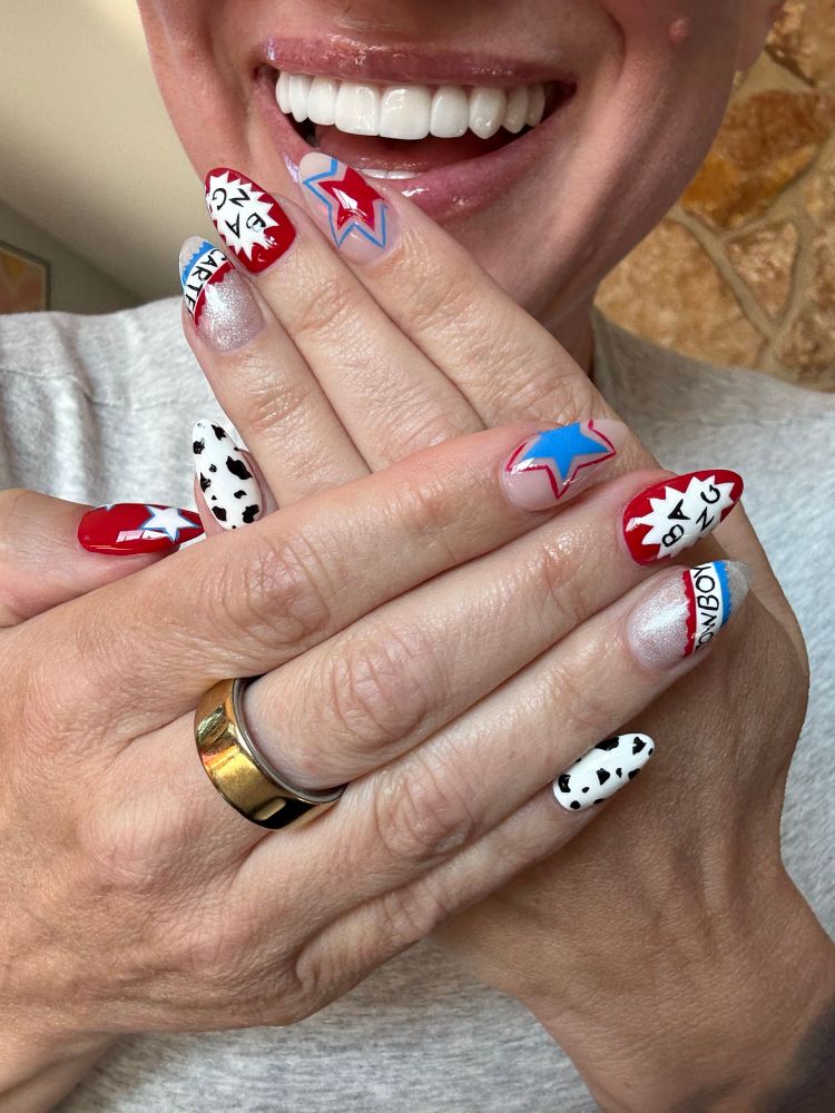 A close-up of hands showing off a playful and bold Beyoncé-themed manicure. Each nail features a unique design, including red, white, and blue stars, comic-style bursts with words like “COWBOY” and “CARTER,” silver shimmer, cow print, and graphic text. The overall look is inspired by Beyoncé’s Cowboy Carter era, blending pop-art flair with western motifs.