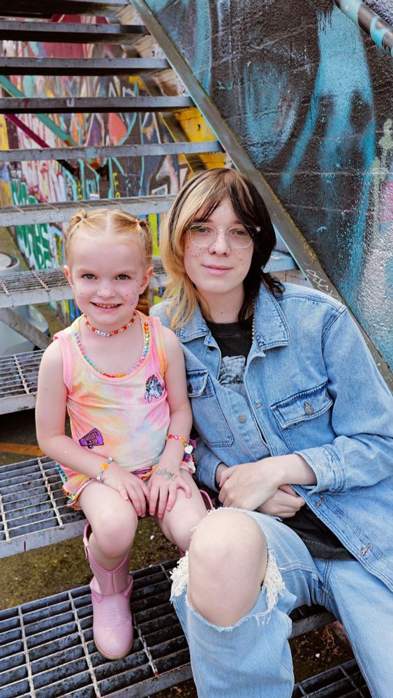 Kris and Marlowe sit together on a metal staircase in front of a vibrant, graffiti-covered wall. Kris wears clear glasses, a denim jacket, and ripped jeans, with rainbow freckles dotted across their face. Marlowe, a young child with braided blonde hair, grins brightly in a tie-dye tank top decorated with playful patches and beaded jewelry. She wears pink cowboy boots and has matching rainbow freckles.