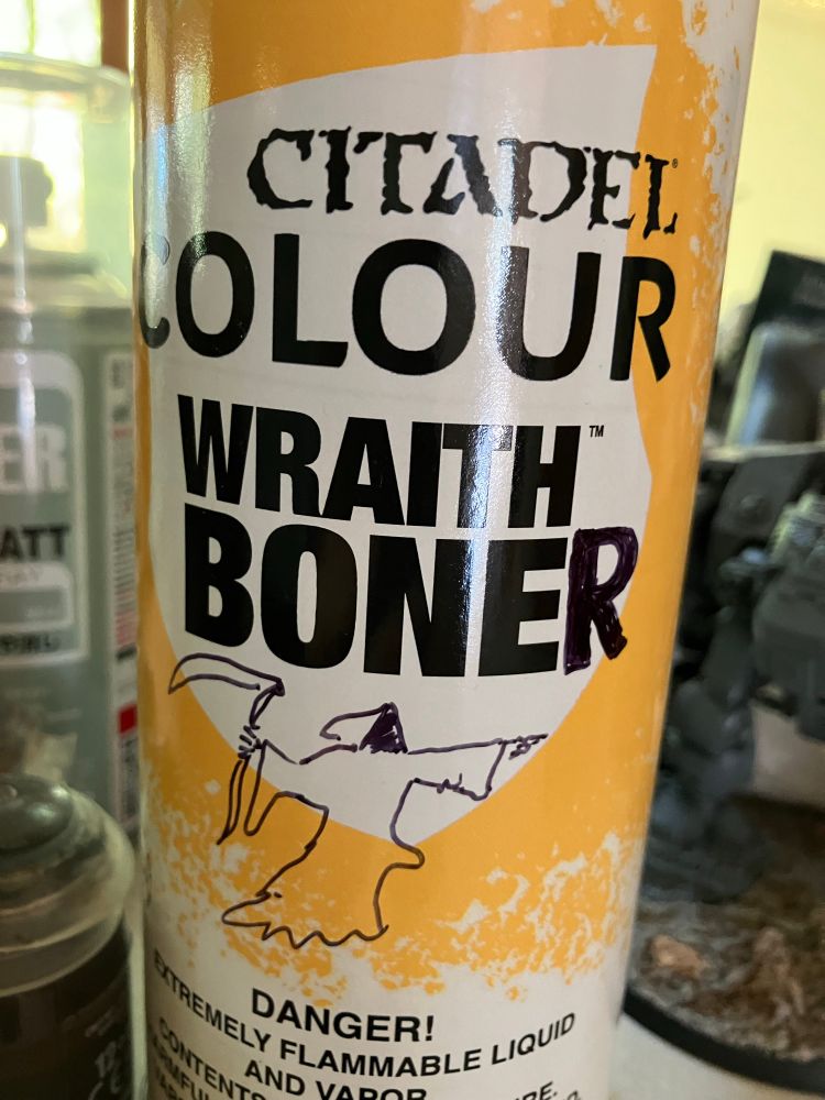 A can of Citadel Spray Primer “Wraith Bone” color, to which an “R” has been added with a sharpie marker as well as a simple illustration of a point long wraith with a scythe and a “telltale protrusion” under his robes on the front.