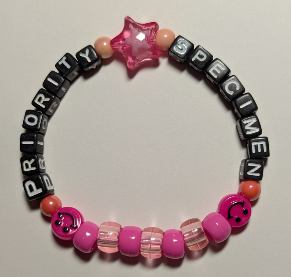 a kandi bracelet that says "priority specimen" in black block with white letter beads, pink smiley faces, a pink star, and pink and light pink pony beads.