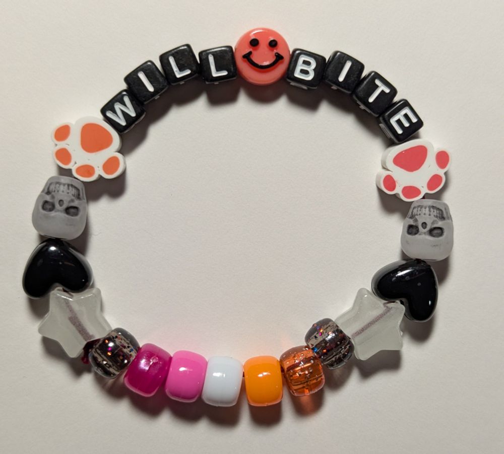 a kandi bracelet that says "will bite" in black block with white letter beads, a pink smiley face, two orange paws, two skulls, two black hearts, two white stars, and lesbian flag color pony beads.