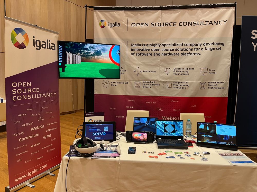 Igalia Booth B8 at OSSSummit EU 2024 including some Igalia banners and a table with multiple computers running demos of Wolvic, Servo, WPE WebKit and graphics. Among them, a Steam Deck and some VR glasses.