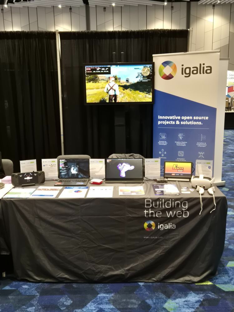 A booth with a tablecloth that says "Building the web - Igalia" atop the table are lots of different devices with demos running on them, from steamdecks to raspberry pis and oculus quest devices. Behind the table a TV projects the XR experience and a sign banner says "Igalia: Innovative open source projects and solutions"