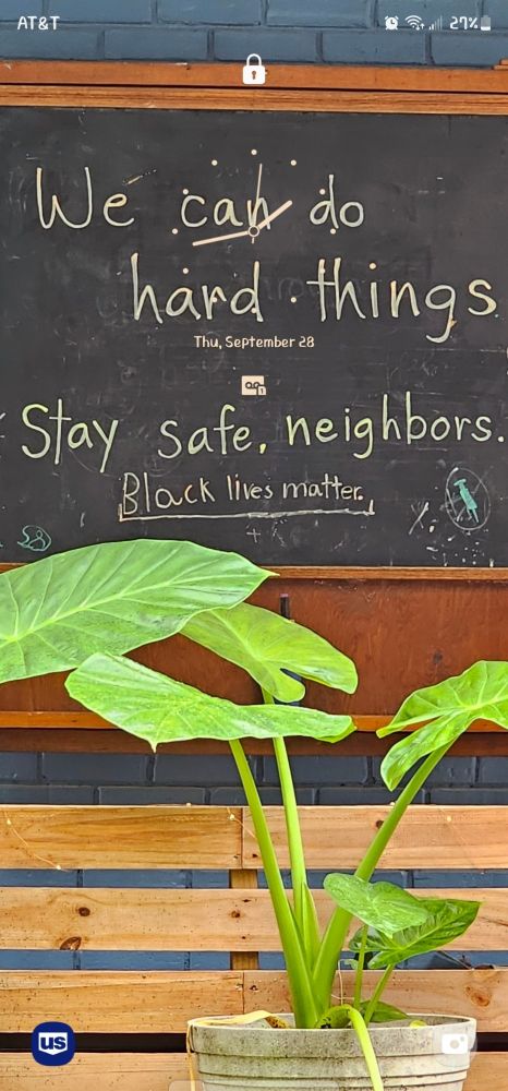 screenshot of a lockscreen with an analog clock showing roughly 1:44 p.m. the photo is a chalkboard on someone's front porch. written on the chalkboard is "we can do hard things ... stay safe neighbors." in the foreground is a wooden plank fence, and in front of that is a large potted elephant ear plant