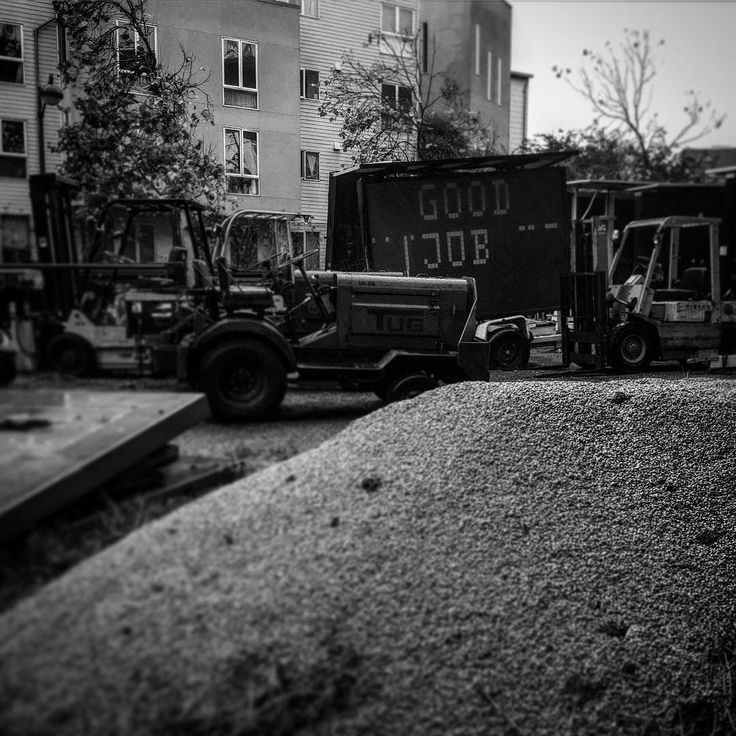 Black and white photo of a construction zone: forklifts and a sign that says "GOOD JOB". Apartment buildings in the background. Large rock filling the lower and right half of the square photo.