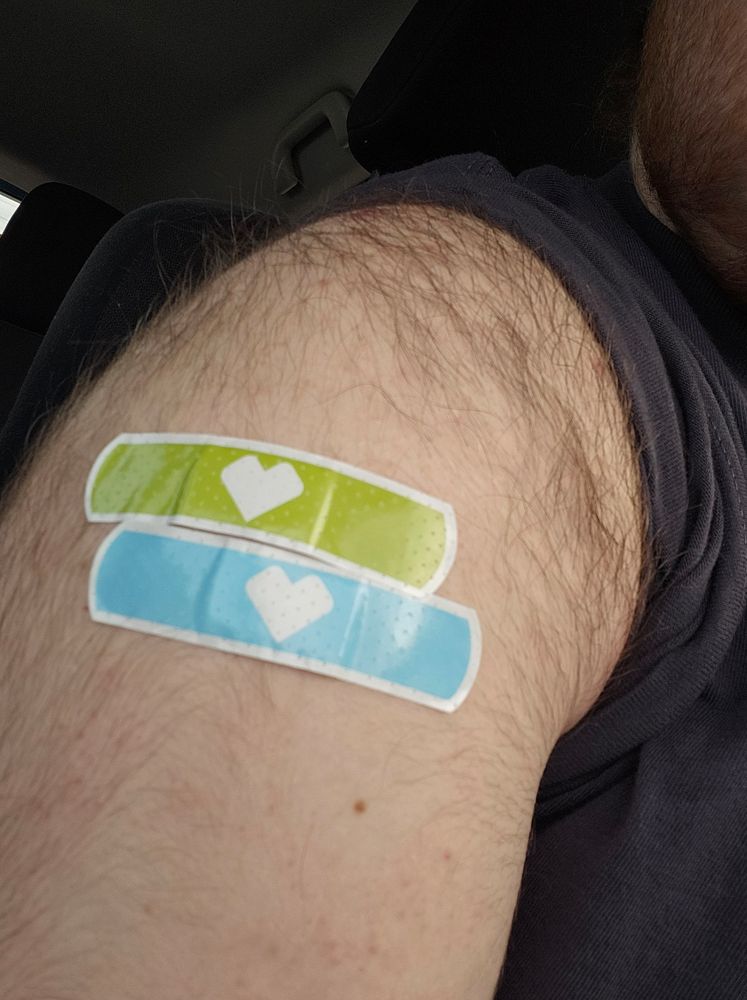 My upper arm with one green band aid sitting right above a blue band aid. One was for a flu shot and the other was for the updated 2025-2026 Covid-19 vaccine.