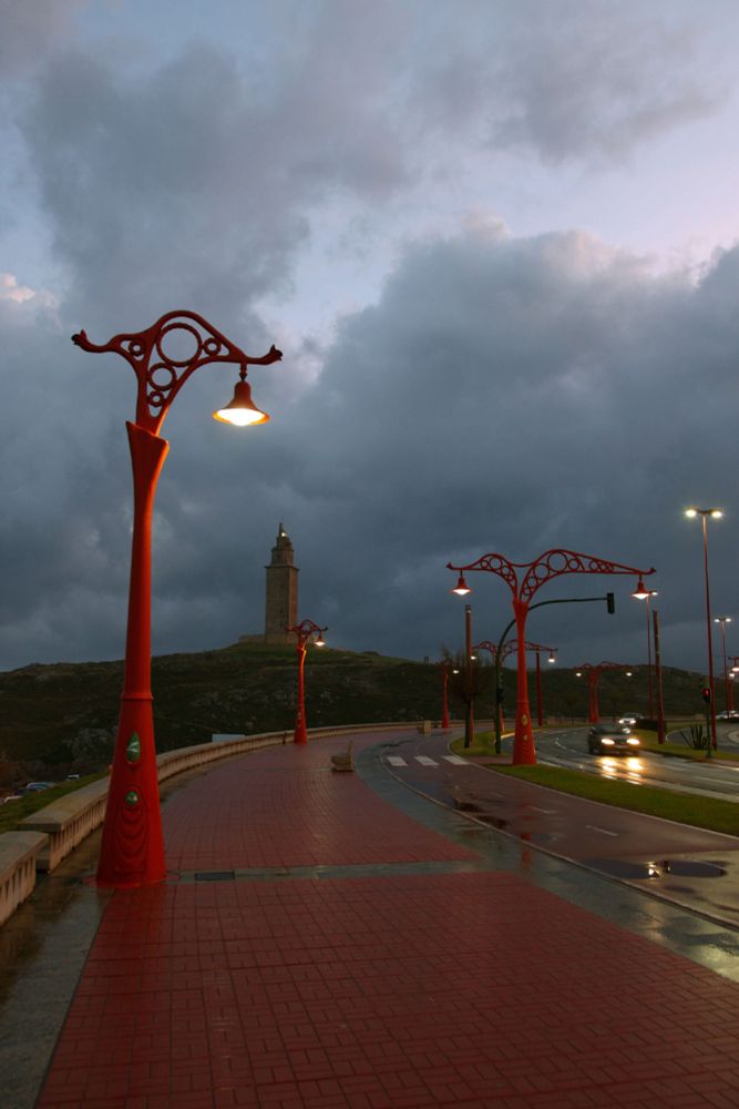 Vista al amanecer (el este queda a la derecha de la imagen y de ahí parece provenir la claridad en un cielo bastante cubierto de nubes) del paseo marítimo coruñés desde, posiblemente, la altura del Aquarium FInisterrae hacia la Torre de Hércoles que se ve al fondo. Las características y artísticas farolas rojas dominan la imagen. Asfalto o zona peatonal húmeda con algo de agua encharcada a la derecha de la imagen. Un par de vehículos vienen de frente al fotógrafo con las luces encendidas, trazando la amplia curva que discurre desde las cocheras del abandonado tranvía turístico. Las farolas aún mantienen sus luces encendidas a pesar de la claridad que empieza a dominar el ambiente.