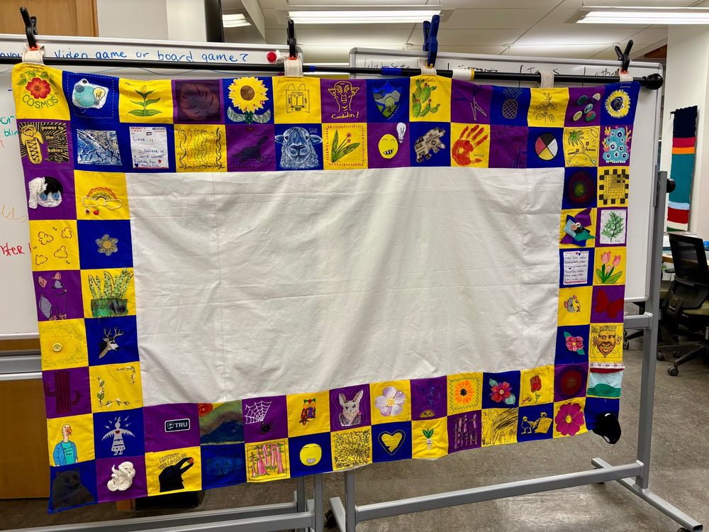 A large fabric quilt hanging on whiteboards. The quilt is made up of many small yellow, blue, and purple squares that are each decorated. The center of the quilt has a large space that is blank. 