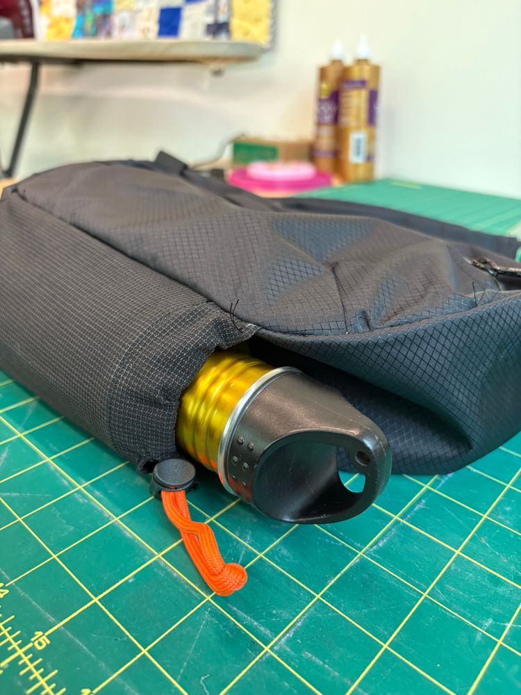A black sling bag on a cutting mat featuring a bottle in a stretchy bottle pocket attached to the bottom