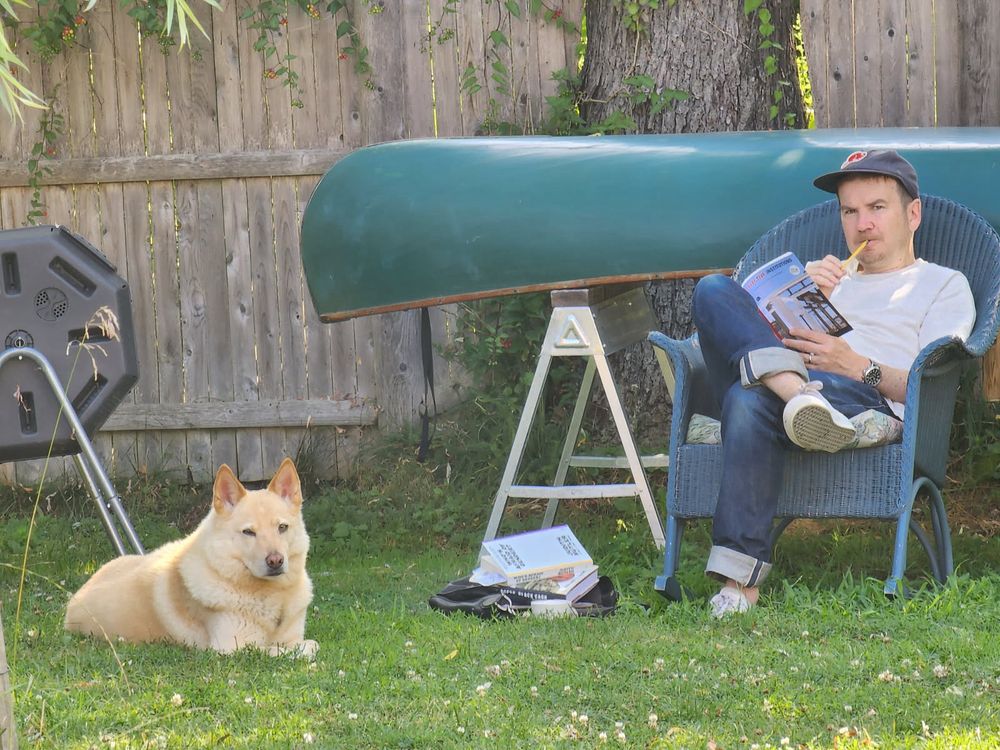 Bento (dog) on ground, me seated and reading. Canoe in background 