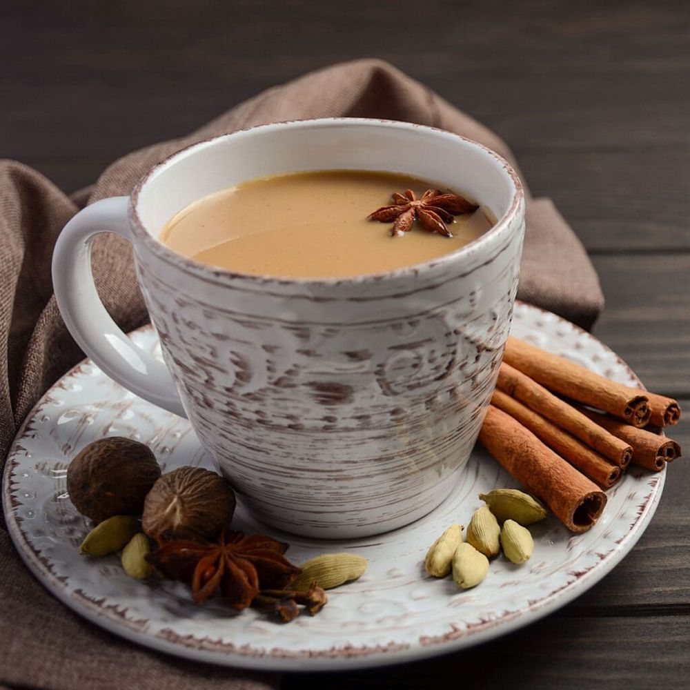 A textured white teacup with various spices arranged around the saucer including cinnamon sticks, aniseed stars, cloves, cardamon pods and nutmeg. The teacup contains chai tea.