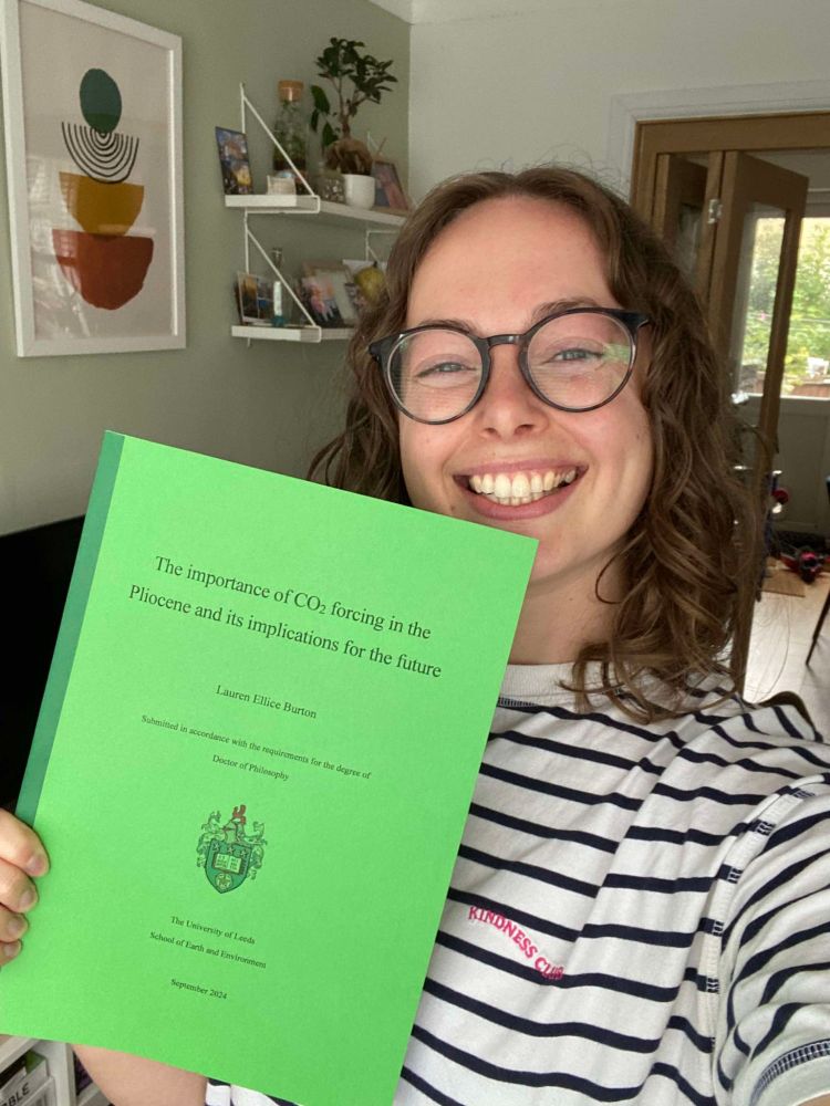 Lauren holding her PhD thesis. Lauren is a white woman with shoulder length brown curly hair and glasses. The thesis has a green cover and is titled "The importance of CO2 forcing in the Pliocene and its implications for the future".