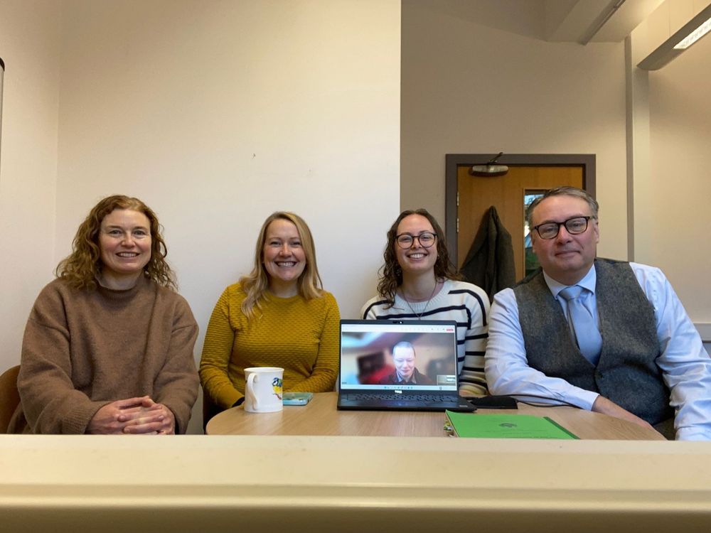 Four people sat at a table smiling towards the camera. A laptop on the table shows a fourth person smiling.