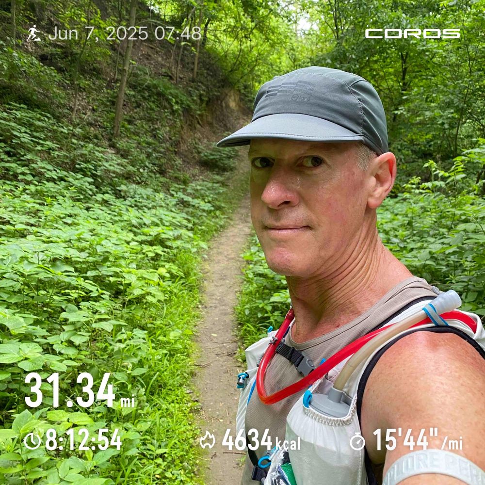 Runner posing with a single track trail and a “gentle” uphill climb in the background. 
