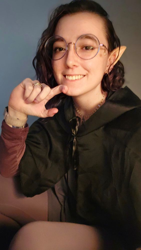 A white woman with brown eyes and brown hair that fades from a mahogany brown to a dark purple. She has on very large circular glasses that have a heart charm on the side. D sneaking with her hand on her chin. She is wearing a cowl and a long gray shirt with a belt around it. Underneath the gray shirt is a purple long sleeve shirt. She is wearing purple leggings. Her ear which is pierced three times also has a fake elf ear on top. The costume was designed to look like Amity from The Owl House. 