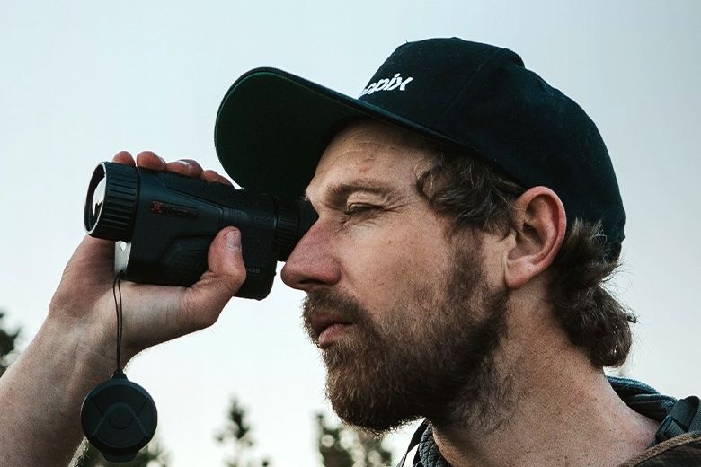 Image (c) Nocpix Lumi thermal monocular image
A side on view of a the head of a white male with dark hair and short beard wearing  black baseball cap and using a palm-sized, black, thermal monocular on his right hand held up to his right eye with his let eye shut.
