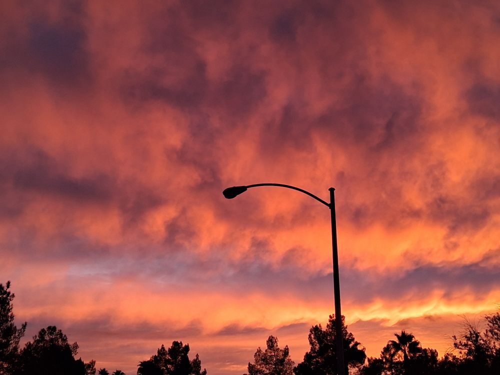 A long street light is silhouette agai st a bright sky full of purple, pink, and peach clouds. 