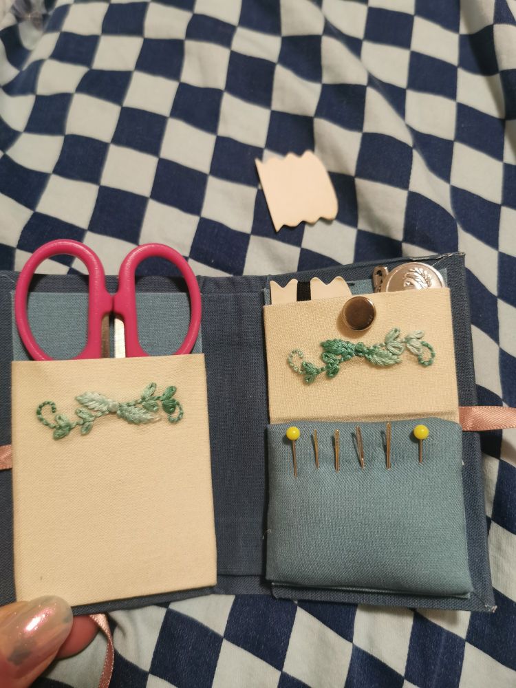Inside of storage box: on the left is pink scissors in their pocket; on the right an embroidery thread card with black thread, magnets (aka cheap needle minders), a safety pin, and a needle threader in the top pocket. The bottom has two pins and four needles stuck in a pin cushion.