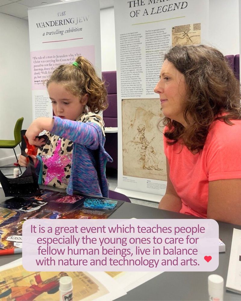 Two people sit at a table working on an art project with black paper and colored markers. A green aloe drink bottle and a pink leaflet are on the table. In the background, others are seated and participating in similar activities. Text overlay reads: “The festival, itself was excellent, there were so many interesting hands on stalls and a great variation.”