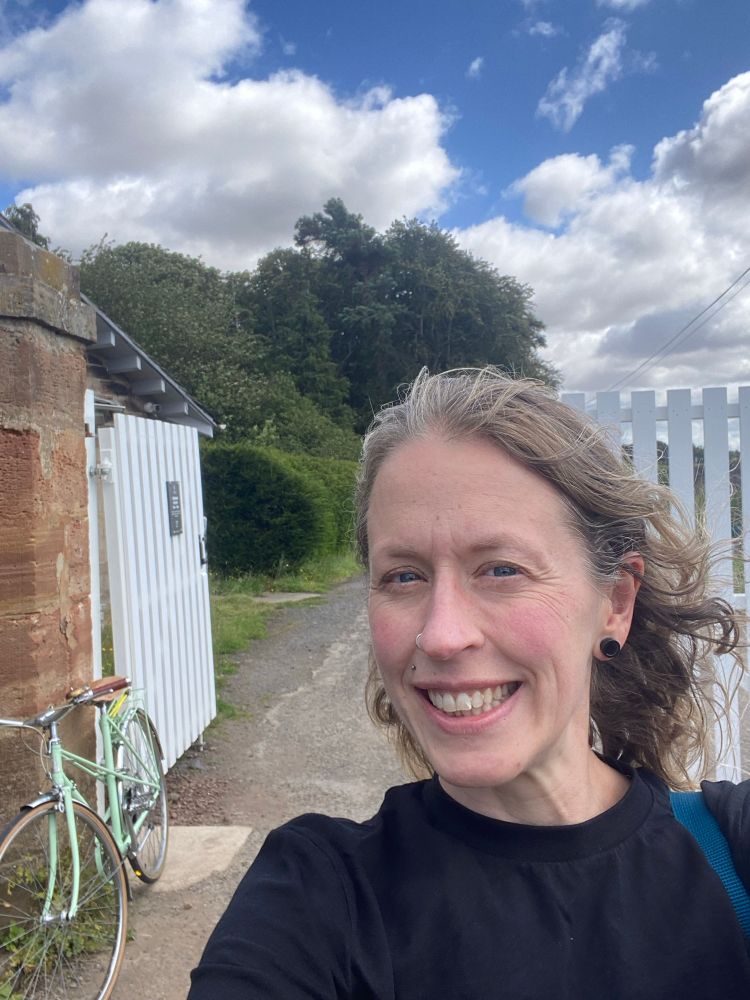 Woman in black smiles at camera in front of an open white wooden gate. Mint green 1980s mixte frame bike leans against wall in background