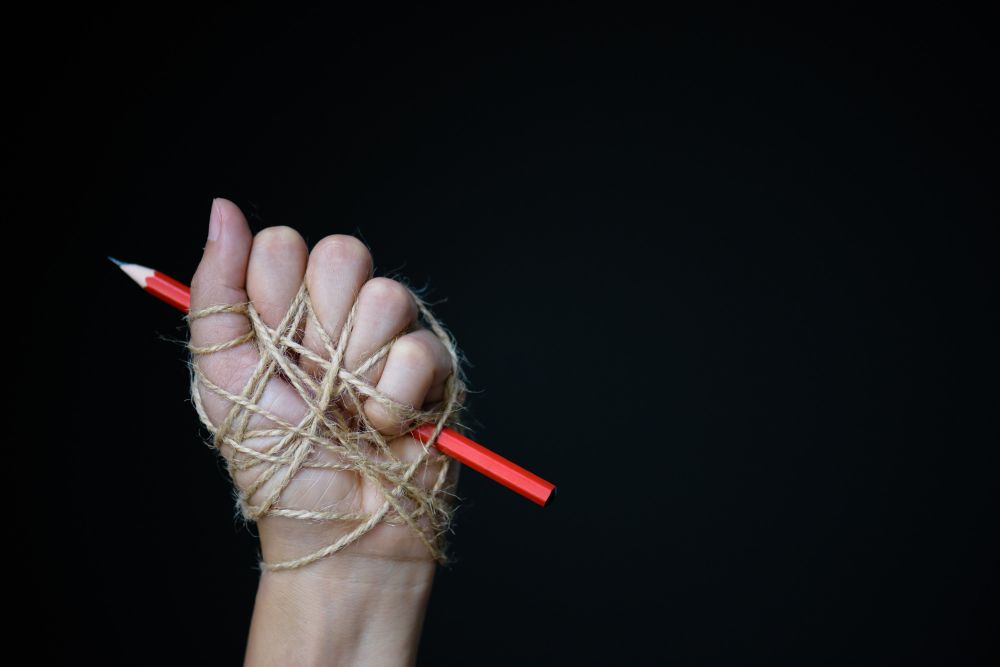 In die Luft gestreckte, mit einem Seil gefesselte Hand. Sie hält einen Bleistift in der Faust. Schwarzer Hintergrund. credits: spukkato @ iStock.