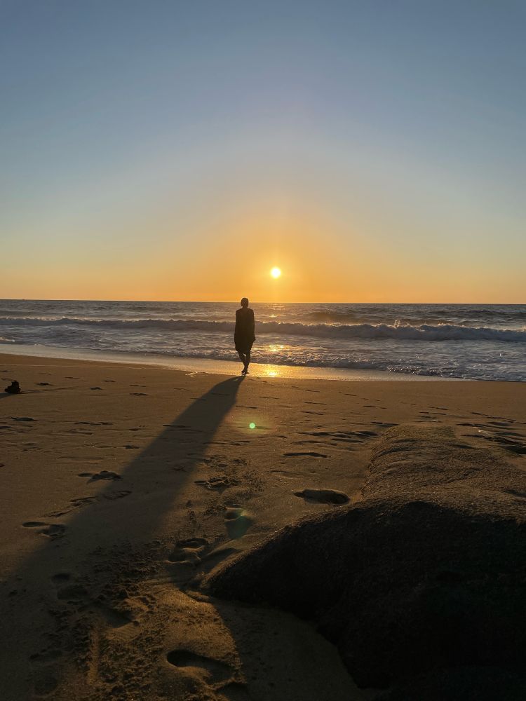 a silhouette of a person walking towards the sea and a setting sun, their shadow reaching back to you across the warm, golden sand
