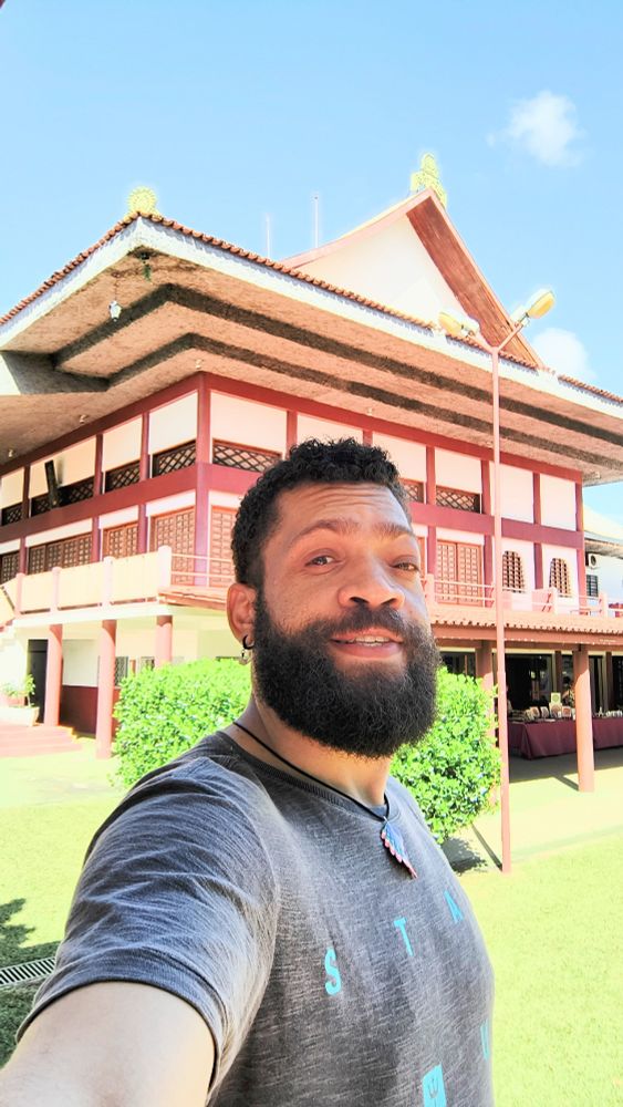 Estou em um ambiente externo, segurando a câmera com o braço esquerdo estendido para tirar uma selfie. Meu rosto está levemente inclinado para a direita, e meus olhos semicerrados por causa da luz intensa. Minha expressão é relaxada, com um leve sorriso. Minha barba é cheia e escura, bem definida, cobrindo toda a região do queixo e bochechas. Meu cabelo é curto, com cachos bem definidos. Uso um brinco pequeno na orelha esquerda e um colar com um pingente colorido.

Minha camiseta é cinza escura, com um efeito desgastado e algumas letras em azul claro estampadas na frente. Meu ombro direito está mais próximo da câmera, criando uma leve perspectiva inclinada na imagem. A luz forte do sol destaca os contornos do meu rosto e o brilho nos meus olhos.

Ao fundo, há um edifício de arquitetura oriental, com um telhado inclinado e ornamentos dourados nas extremidades. A estrutura combina tons de vermelho e branco, com pilares e janelas de madeira decoradas com padrões vazados. A parte inferior do prédio tem um espaço coberto, onde mesas estão organizadas com objetos expostos.

O ambiente ao redor é composto por um gramado bem cuidado e arbustos verdes, contrastando com as cores quentes do edifício. À direita, um poste de iluminação avermelhado se inclina ligeiramente para dentro da imagem. O céu está azul e sem nuvens significativas, e a luz intensa do sol cria sombras nítidas no cenário.

