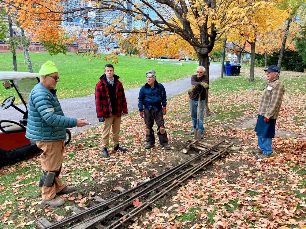 A group of TRHA volunteers and our museum curator stand beside one of the pathways in an autumnal-looking Roundhouse Park, discussing a disconnected miniature railway switch sitting on the ground between them.