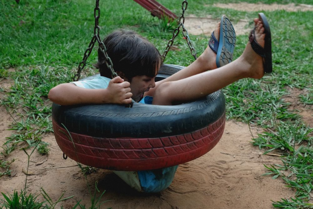 Parque infantil gramado, foco em uma criança em balanço de pneu preto e vermelho, preso por correntes. A criança está caindo no espaço interno do pneu e atingindo o solo. Ela segura firme nas correntes. Seus pés estão para o alto, calçados com chinelo de dedo azul.