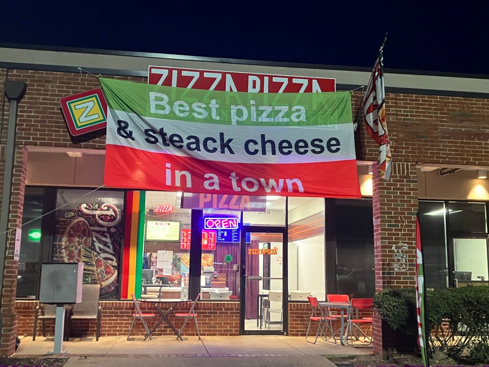 brick restaurant storefront for zizza pizza, its sign covered up by a banner with three vertical color stripes, green white red, to match the Italian flag: 
Best pizza (green)
& steack cheese (white)
in a town (red)