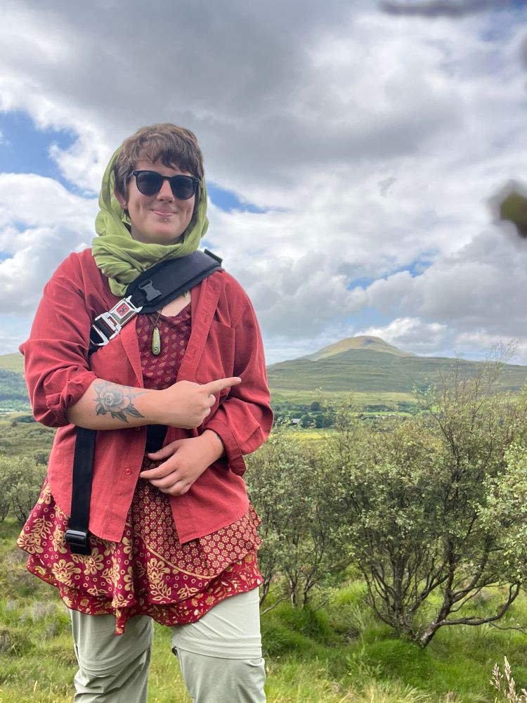 A photo of a person standing in an open moorland pointing to a hill in the distance.