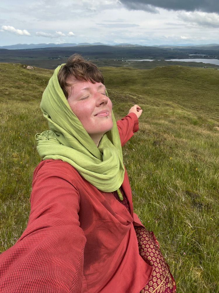 A photo of a person closing their eyes and smiling on a green hill. 