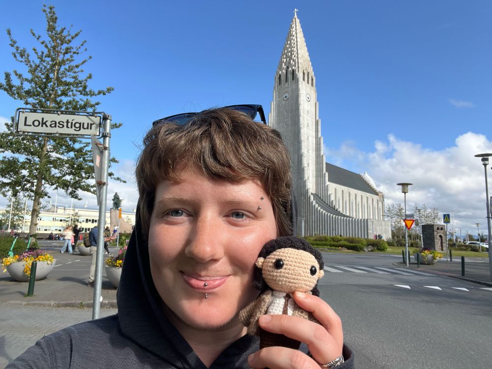 A selfie of a person holding a small crochet doll of Loki from the 2021 TV series in front of a large white church and a street sign that reads “Lokastígur.”