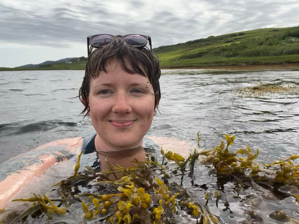 A photo of a person smiling submerged in dark water with sea kelp.