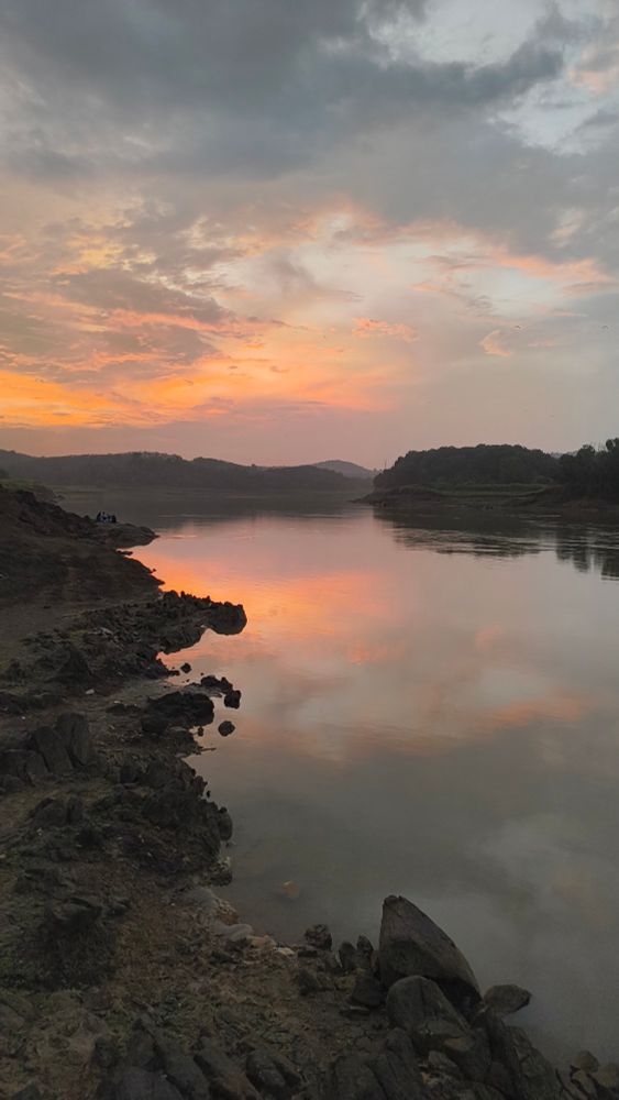 Sky at sunset with orange clouds. The clouds are reflecting onto a river with rocky banks. Small hills appear at the horizon. 
