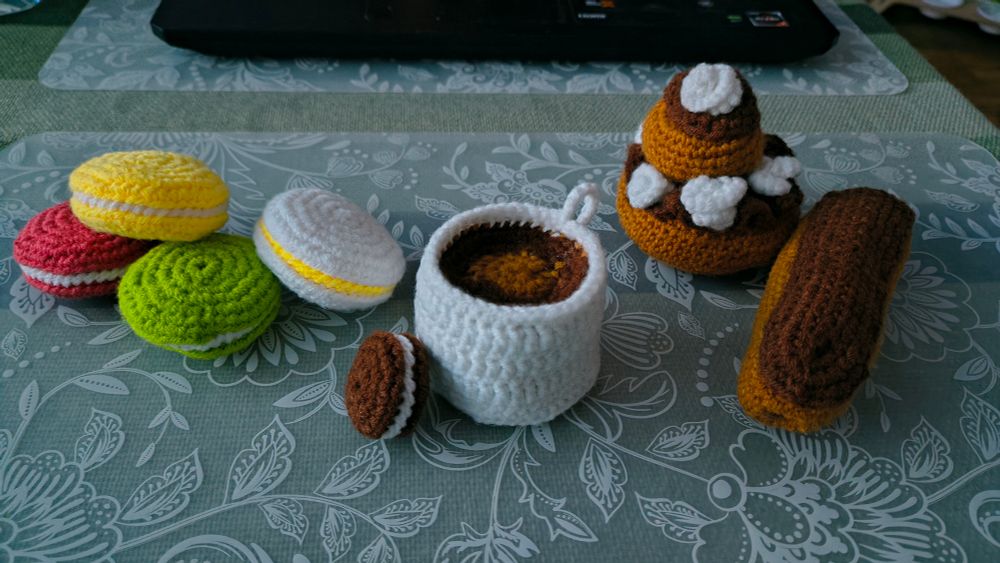 Photo de groupe de mes Amigurumi de pâtisseries. Il y a 4 macarons, un petit biscuit, une tasse de chocolat ou café selon l'envie, une religieuse au chocolat et un éclair au chocolat.