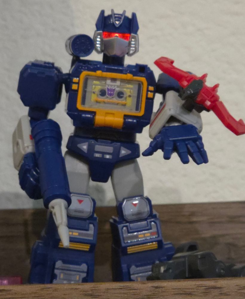 Photo of a Soundwave figure. His visor glows. Laserbeak sits on his left arm. Ravage just peaks over a wood ledge in the foreground. 