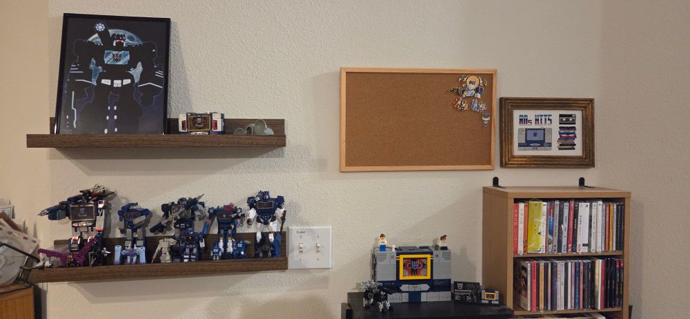 Picture of a wall that has a couple shelves that have various Soundwave toys and a framed print, CD shelving where a LEGO Soundwave sits, a corkboard with some pins, and the framed cross stitch piece. 
