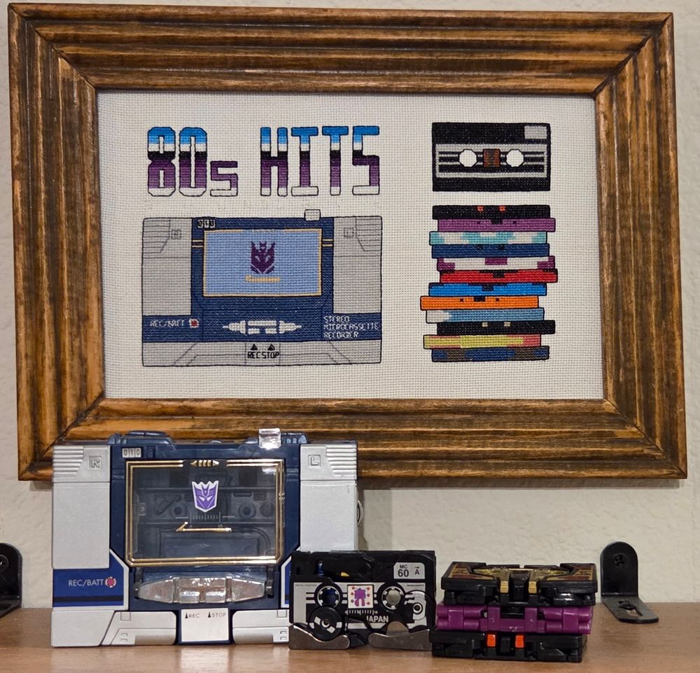 In the foreground sits toys for Soundwave, Ravage, Laserbeak, Rabat, and Buzzsaw, all in alt modes, the last three stacked next to the other two. Behind them is a hung cross stitch that contains the aforementioned characters plus many more stacked cassettes and the words "80s HITS". 