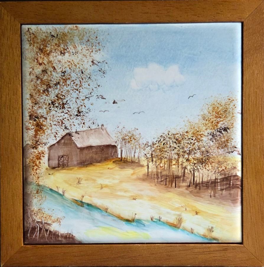 Painted tile set in wooden frame trivet. A brown barn sits in a yellow-brown field, a patch of trees sitting to the right. In the foreground, a creek runs across with a tree on the left side of the tile. The sky is blue with simple bird silhouettes and a couple of white clouds. 