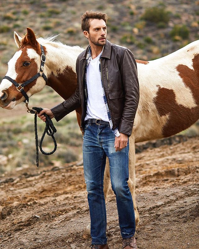 Colour photo of Brazilian man outside leading a horse, cropped to full body and front of horse. The man is wearing blue jeans, white t-shirt, blue plaid, and a brown leather jacket and boots. The horse is a skwebald of chestnut and white being led with a grey headcollar and leadrope. They are walking to the left but he is looking off to the right with a neutral expression.